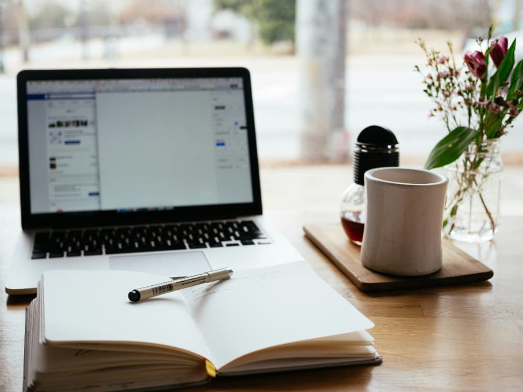 Aufgeschlagenes Notizbuch mit Stift, Laptop und Kaffee auf einem Schreibtisch am Fenster